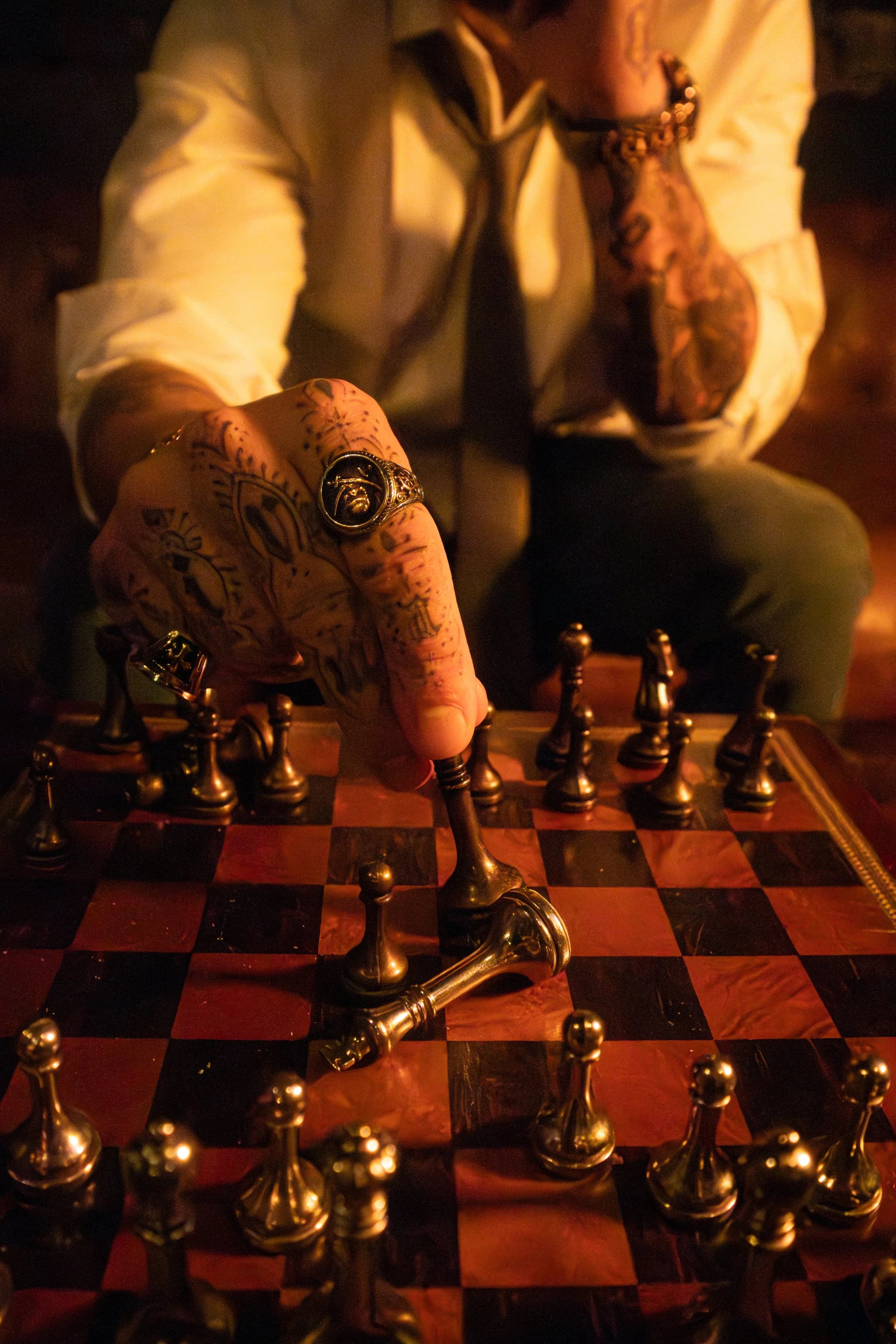 Tattooed hand wearing a signet ring moving a brass chess piece across a lit board